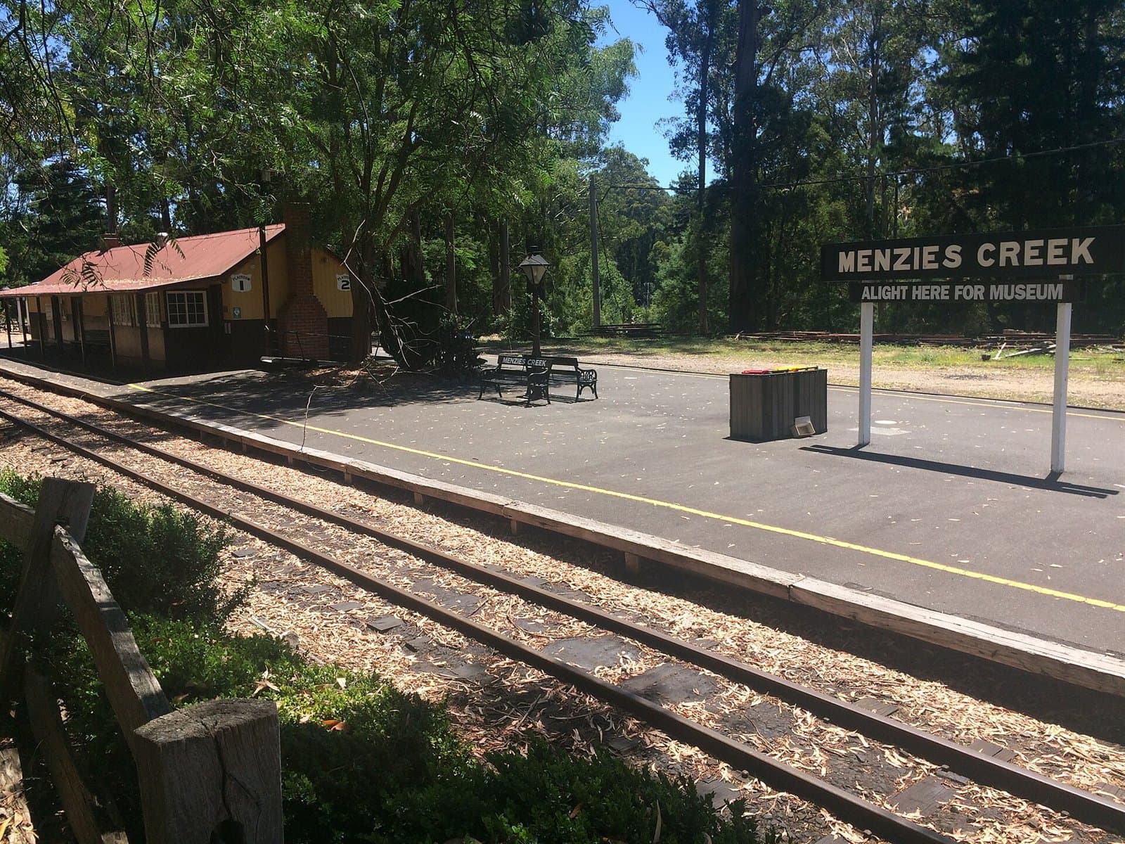 An empty railway platform with a station sign and a timber building under tall trees