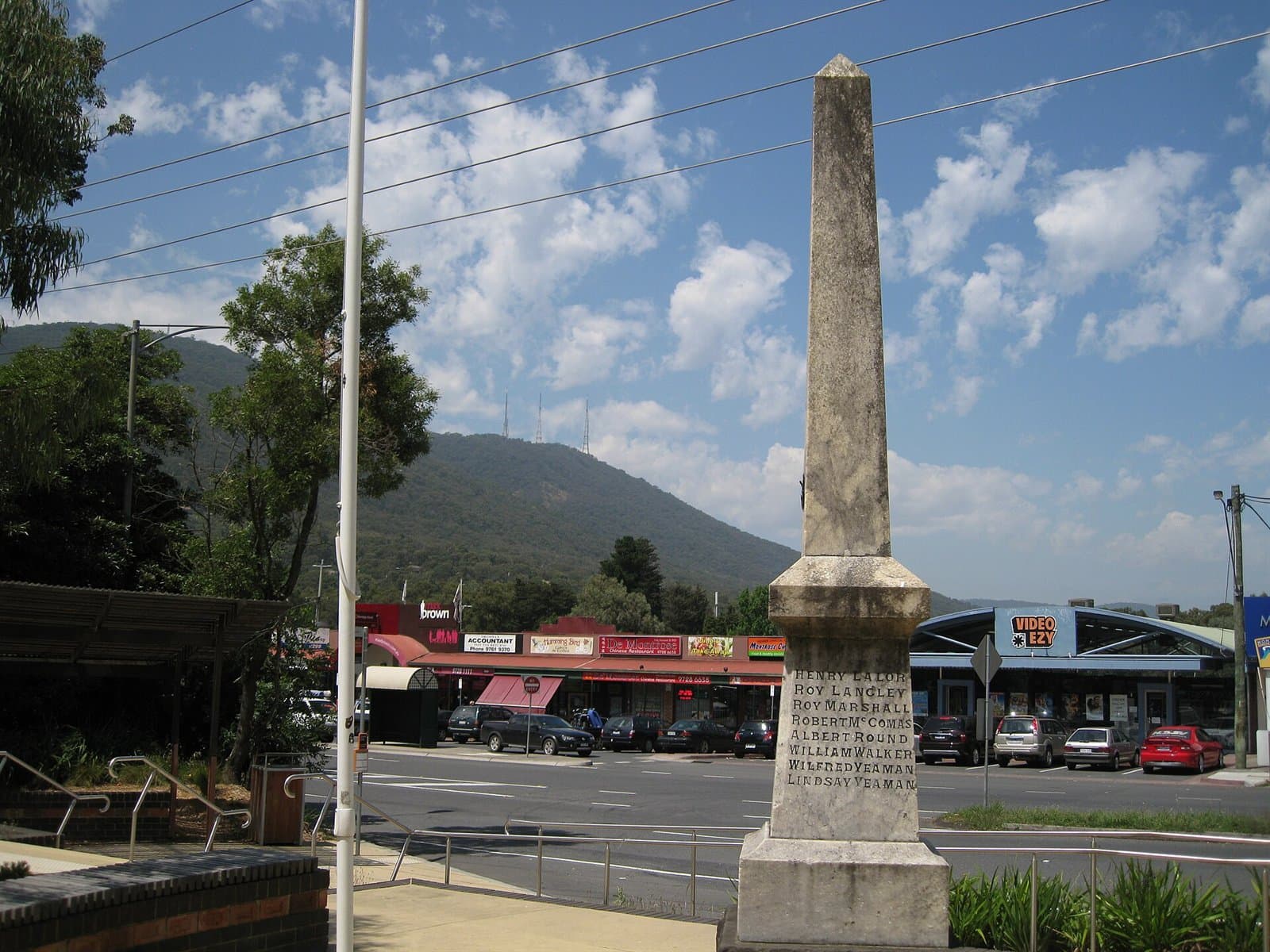 A stone memorial obelisk at a road junction, with shops and Mount Dandenong behind